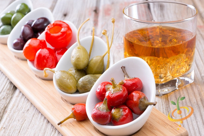 Assorted pickled peppers and olives with a glass of amber beverage on a wooden serving board